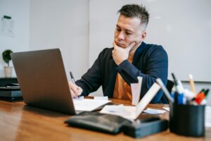 Concentrated man working on laptop and taking notes at a wooden desk in a home office.