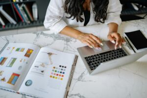 A woman works on a laptop at a marble desk with a design book and notebook nearby.